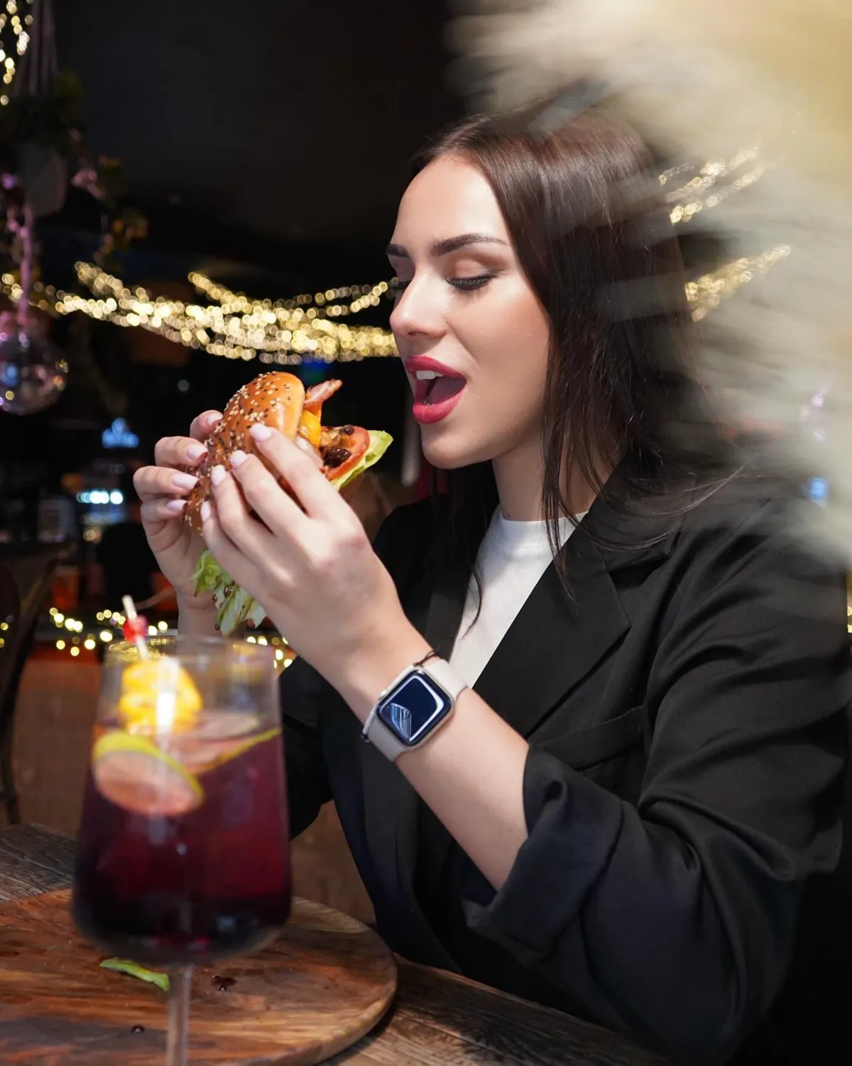 Viese kafejnīcā ar burgeru rokās un dzērienu uz galda, fonā svētku gaismiņas