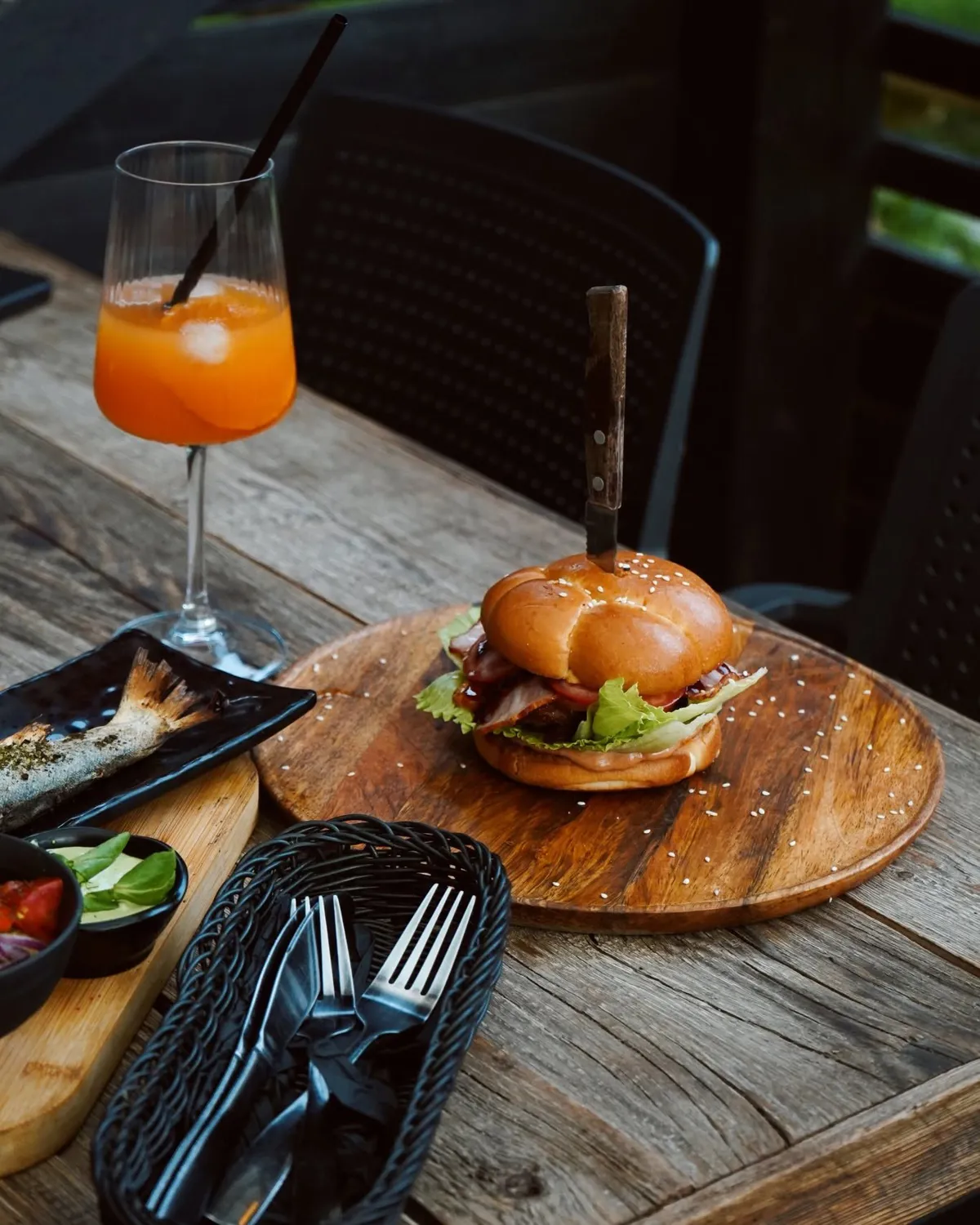 Burgers ar bekonu uz koka dēlīša, glāze ar oranžu dzērienu un cepta forele uz terases galda